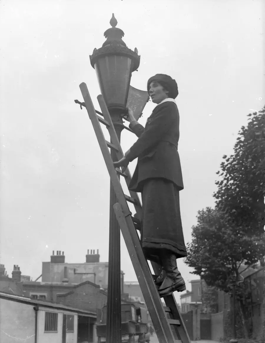 A woman in vintage clothing stands on a ladder lighting a street lamp. Buildings and trees are visible in the background.