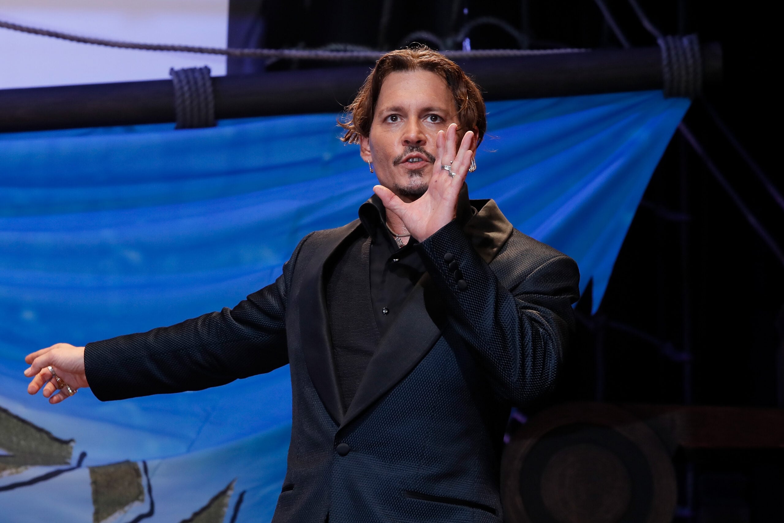 A man in a dark suit gestures with his left hand raised, standing in front of a blue, nautical-themed background.