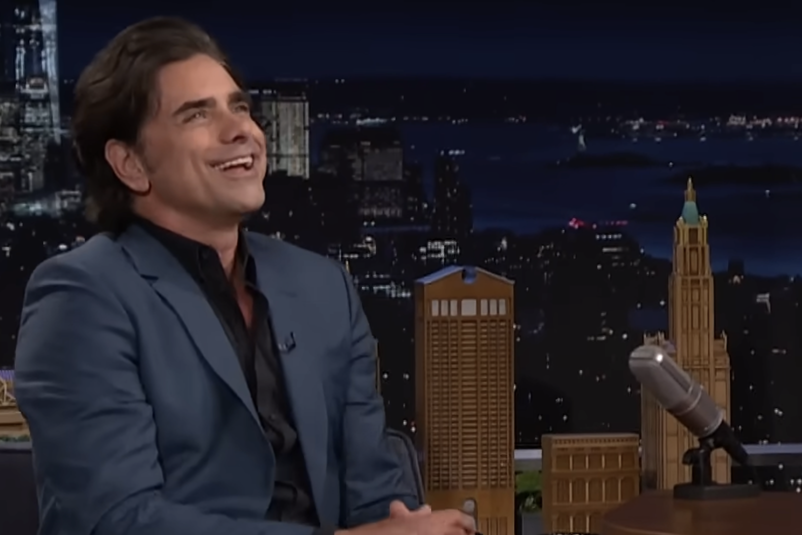 A man in a blue suit sits and smiles on a talk show set at night, with a microphone and miniature city buildings in front of him and a cityscape background visible behind.