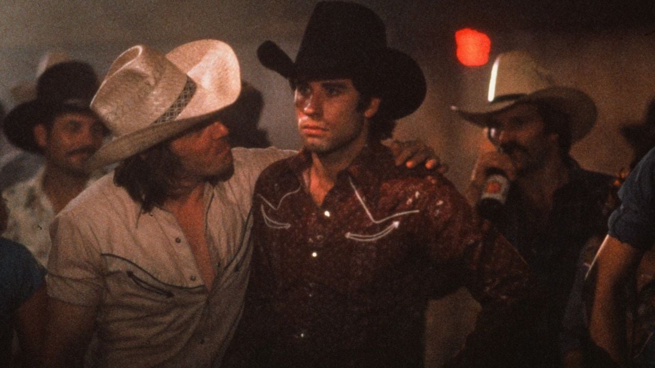 Two men in cowboy hats stand close in a crowded, dimly lit bar. One man comforts the other, who looks serious. Other people in cowboy hats are visible in the smoky background.