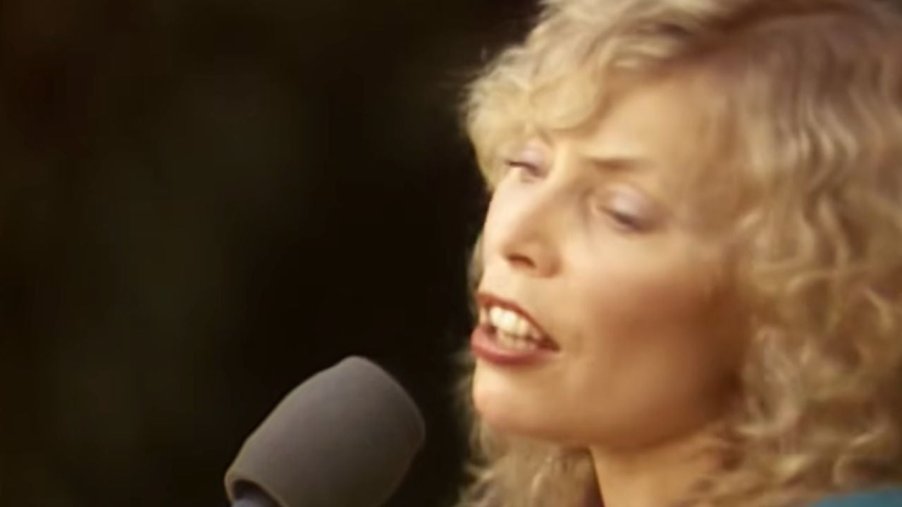 A person with curly blonde hair sings into a microphone, eyes gently closed, with a blurred dark background.