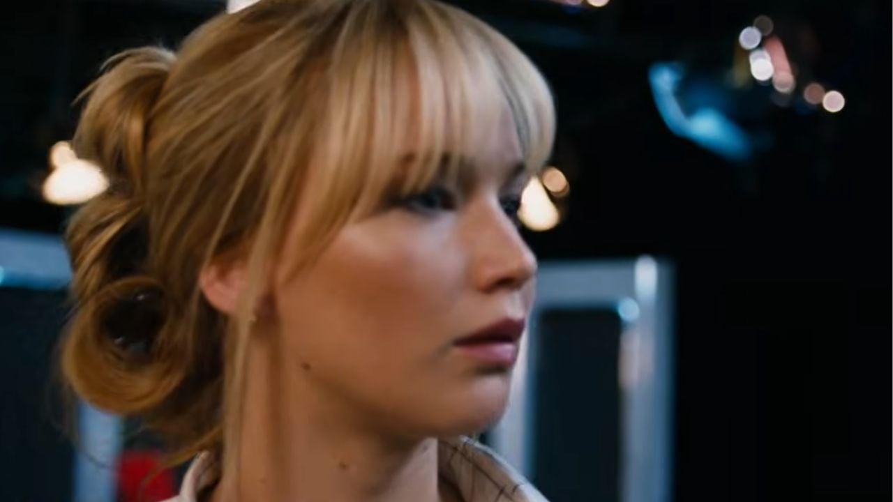 A woman with blonde hair styled in a loose updo and wearing a striped shirt looks to the side in a softly lit indoor setting with blurred lights in the background.