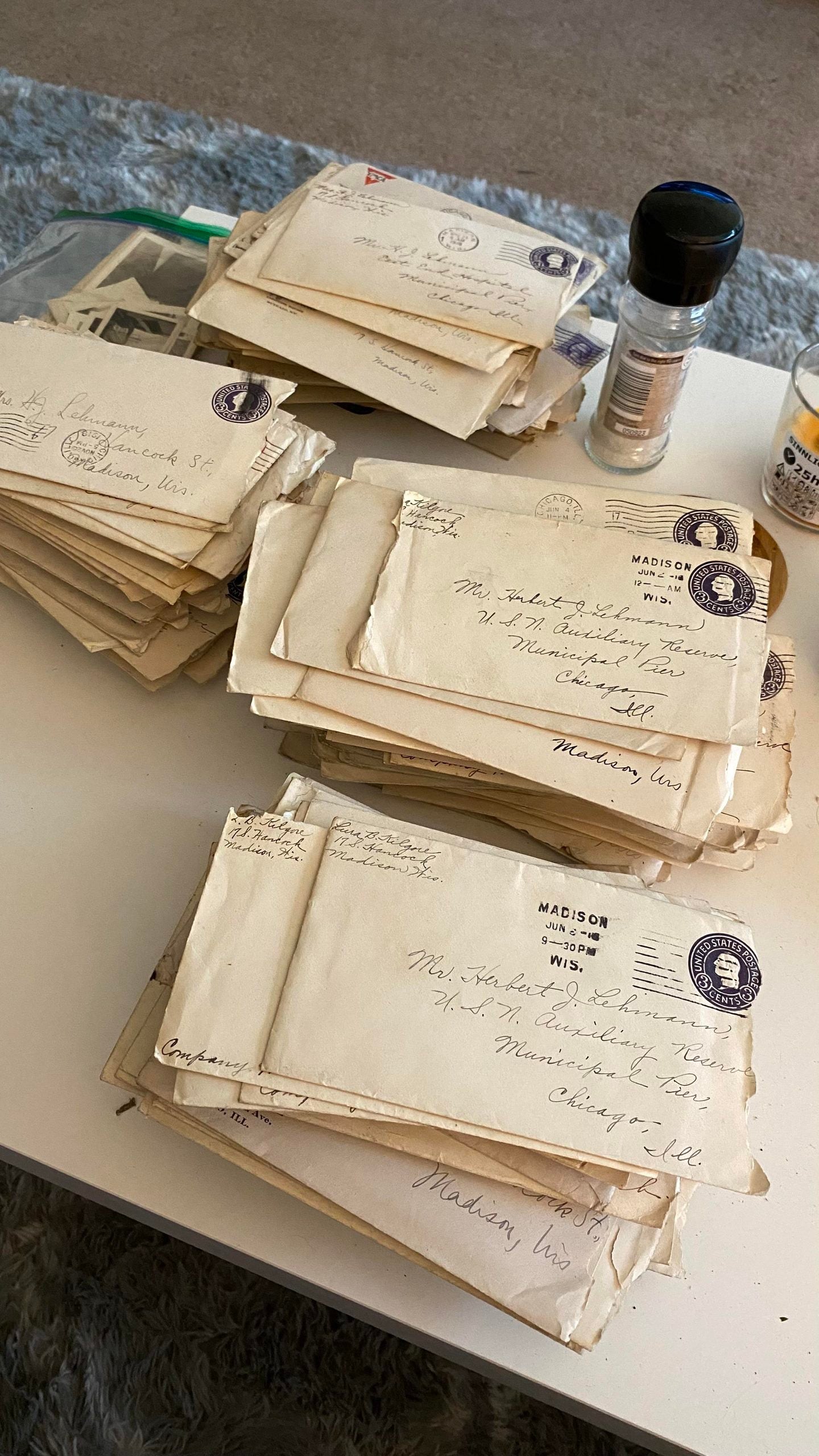 Stacks of old, yellowed envelopes and letters with handwritten addresses sit on a white table, alongside some black-and-white photos and a small spice jar.