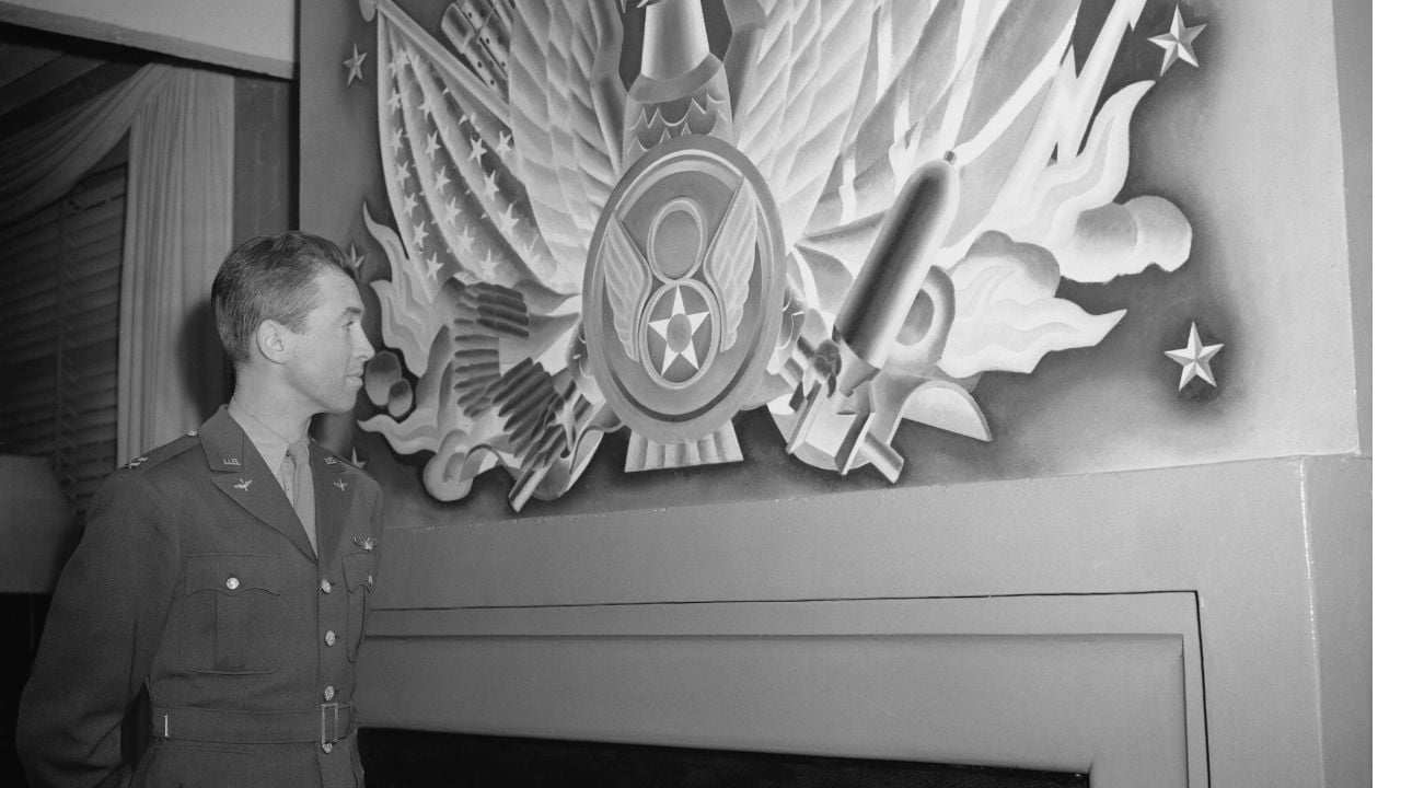 A man in a military uniform stands looking at a large, detailed mural featuring an eagle, stars, flags, and symbols, including a radioactive sign and flames.