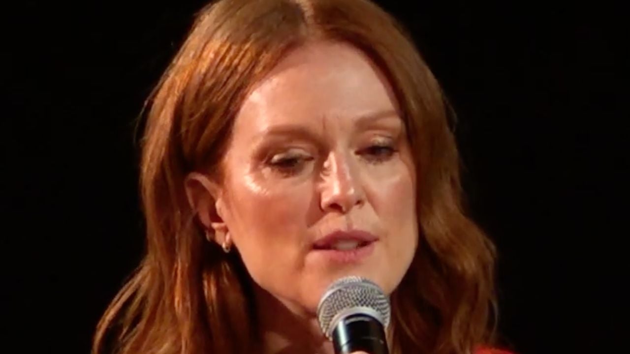 Woman with wavy auburn hair speaks into a microphone against a dark background, appearing thoughtful and focused.