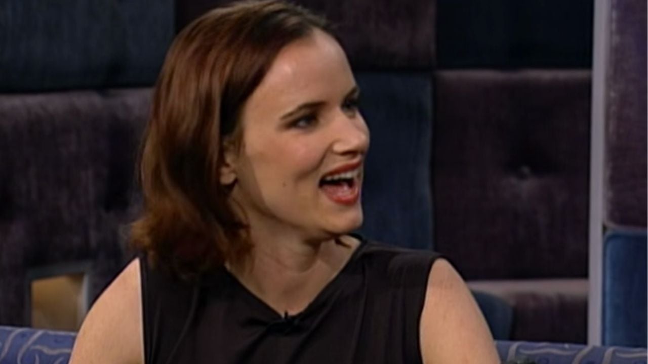 A woman with short brown hair wearing a sleeveless black dress smiles while speaking, seated on a couch with a dark, patterned background.