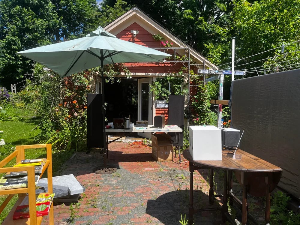 A sunny backyard with a red shed, tables, chairs, a large umbrella, books, and audio equipment on display, surrounded by lush green plants and trees.