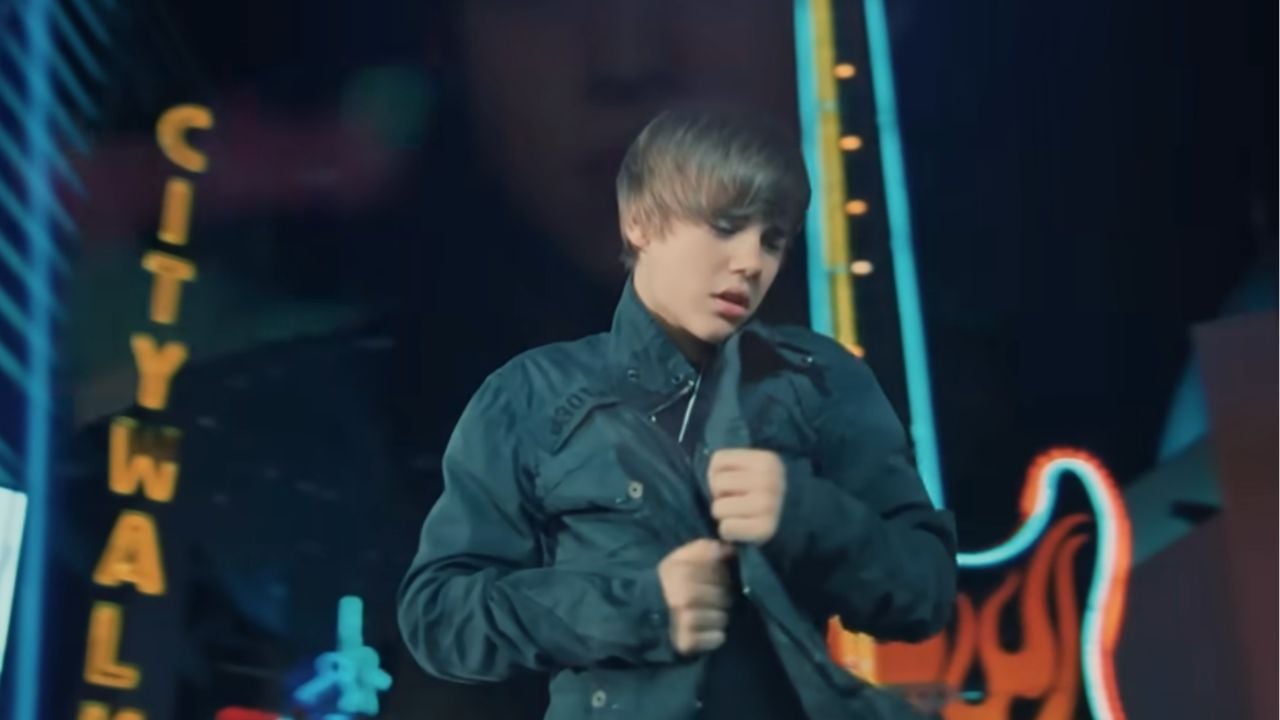 A young person with brown hair stands outdoors at night, adjusting their jacket. Bright neon signs and a guitar-shaped light can be seen in the blurred background, indicating an urban entertainment area.