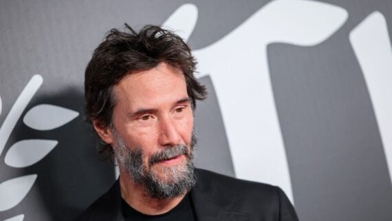 A man with a salt-and-pepper beard and tousled dark hair stands in front of a gray background with large white text and a laurel design. He wears a black jacket over a black shirt and has a contemplative expression.