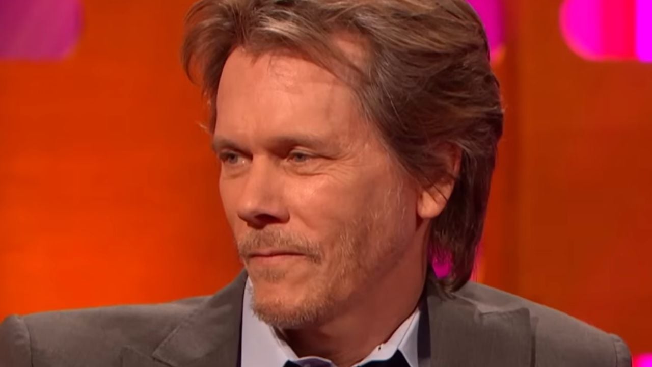 A man with light brown hair and a trimmed beard, wearing a suit and light blue shirt, sits in front of a vibrant orange and pink background, looking slightly to the side with a subtle smile.