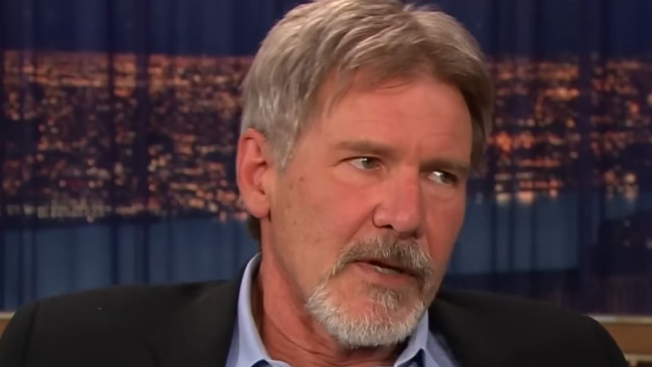 A man with gray hair and a beard, wearing a suit jacket and blue shirt, is speaking with a serious expression. The background is blurred city lights at night.