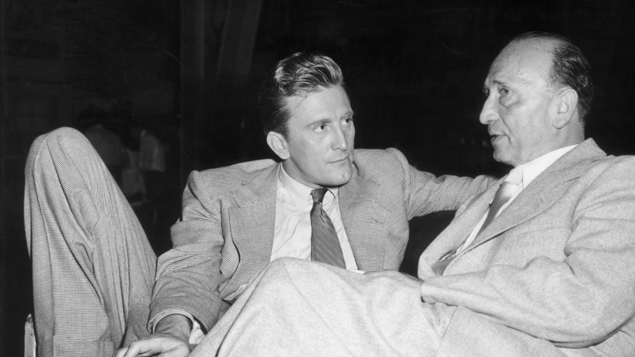Two men in suits sit close together, engaged in conversation. One man has his arm around the back of the couch, and both appear serious and focused. The photo is black and white, with a dark background.