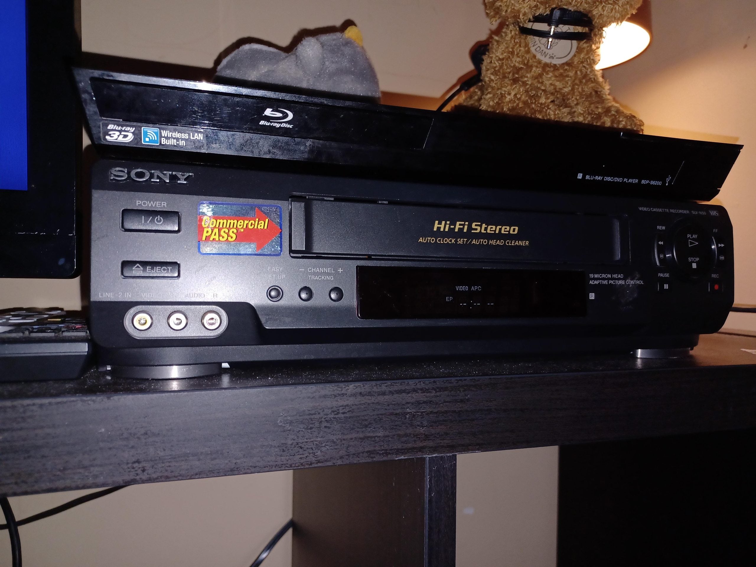 A Sony Hi-Fi Stereo VCR sits on a dark shelf, with a Blu-ray player and stuffed animal on top. The VCR has a “Commercial PASS” sticker and several input/output ports visible on the front panel.