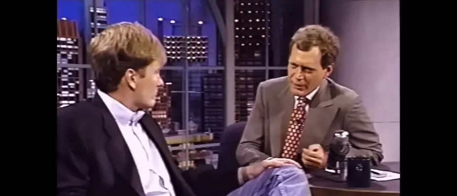 Two men talk on a late-night talk show set; one wears a black blazer and jeans, the other a gray suit with a patterned tie. City lights are visible through large windows behind them.