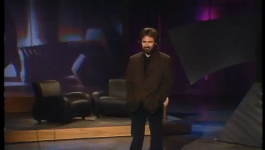 A man with a beard stands on a dimly lit stage with his hands in his pockets. Behind him are two black armchairs and abstract set pieces, illuminated by purple and blue lights.