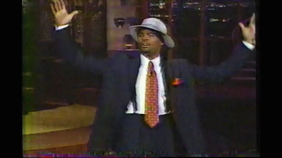 A man in a suit and hat stands indoors with his arms raised. He wears a white shirt, patterned tie, and pocket square, and appears to be speaking or performing.