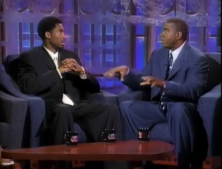 Two men in suits sit on a talk show set, engaged in conversation, with coffee mugs on a table between them and a brightly lit, geometric background behind them.