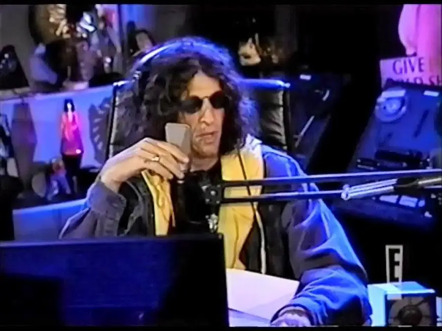 Man with long curly hair and sunglasses speaks into a microphone in a radio studio, surrounded by equipment and decorations.