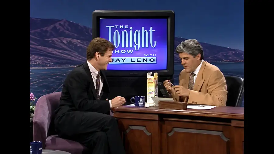 Two men sit at a talk show desk. One man in a suit laughs while the host, seated at the desk, examines a small object. A TV behind them displays “The Tonight Show with Jay Leno.”