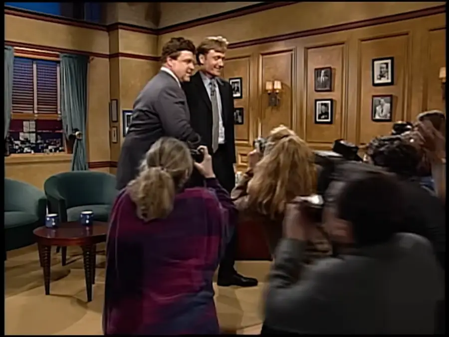 Two men in suits pose together on a TV talk show set as a group of photographers take pictures. The set includes chairs, a table, and framed photos on wood-paneled walls.