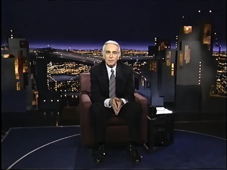 A man in a suit sits on a chair in a TV studio with a nighttime cityscape backdrop, hands clasped, facing the camera.
