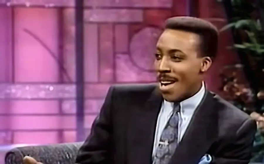 A man in a suit and tie sits on a talk show set with pink and purple lighting, smiling and conversing.