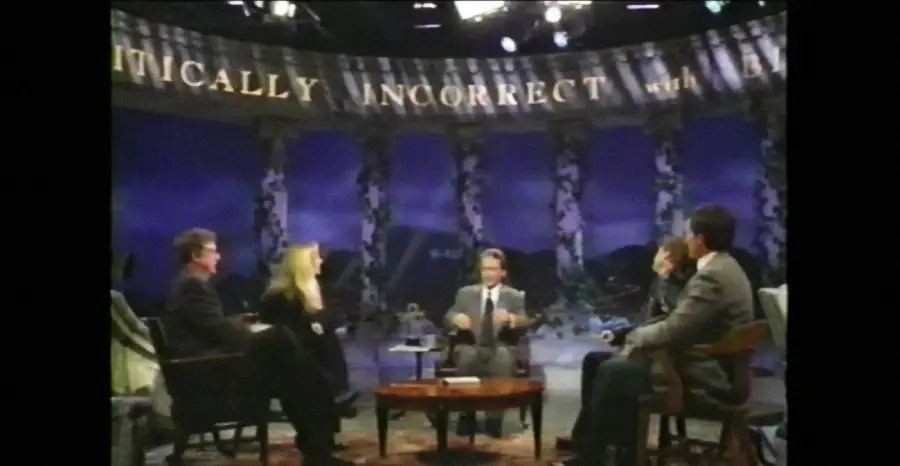 Five people sit in a semicircle on a TV set with “Politically Incorrect” written behind them. The host sits in the center, gesturing, while panelists listen. The background is blue with decorative columns.