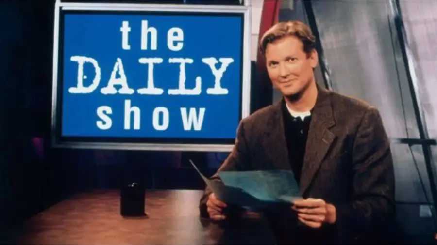 A man in a suit sits at a desk holding papers, with a large screen behind him displaying “the DAILY show” in white text on a blue background.