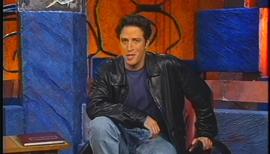 Man with dark hair sits casually in a chair, wearing a black leather jacket and jeans. The colorful background has blue and orange abstract patterns, and there is a book on a table to his left.