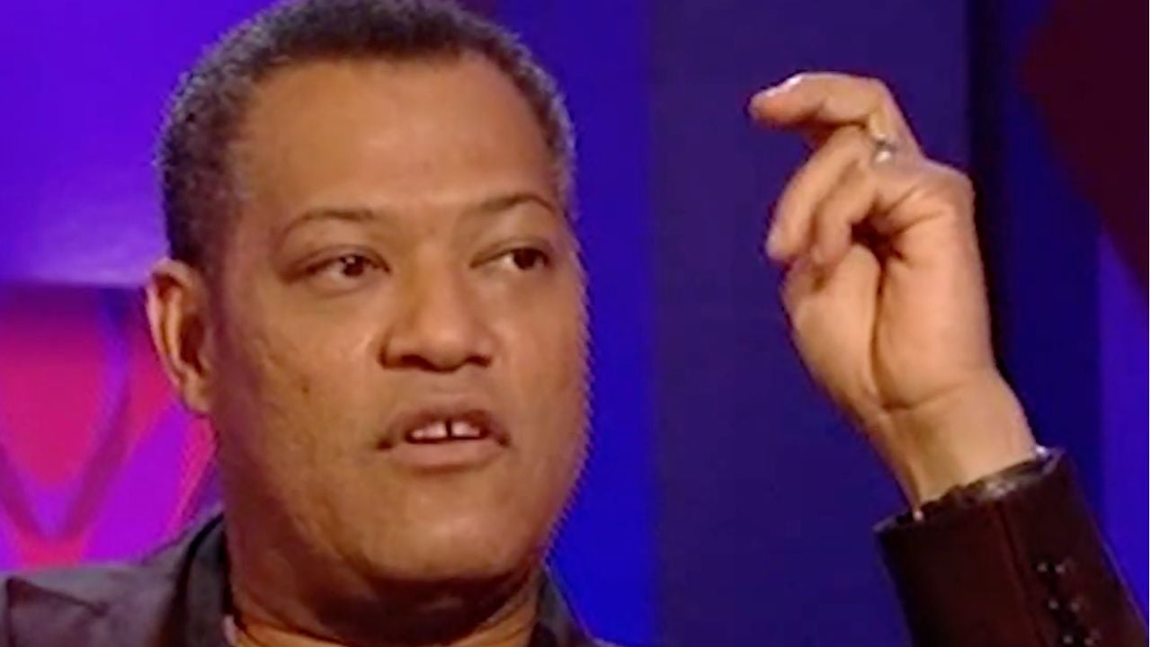 A man with short hair is sitting indoors, holding his hand up near his face as if gesturing while speaking. The background is purple and pink.