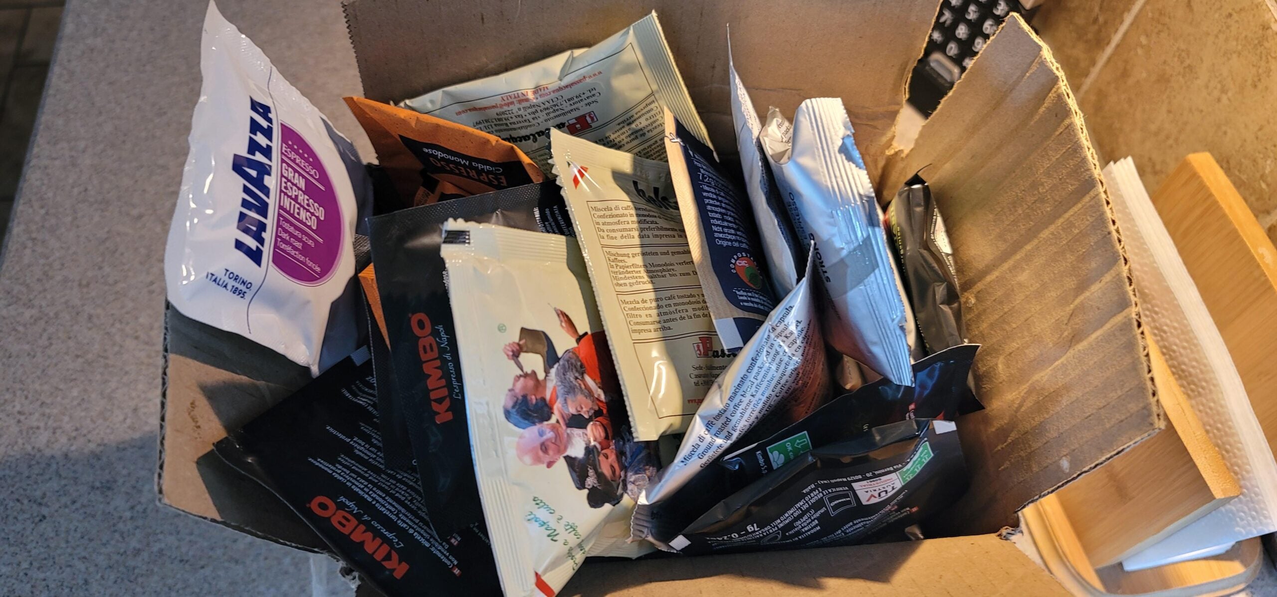 An open cardboard box filled with various brands of small coffee sample bags, including Lavazza and Kimbo, sits on a kitchen counter next to a wooden napkin holder.