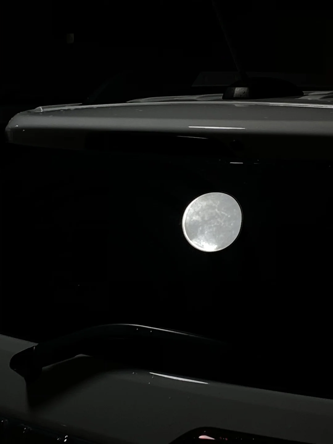 A bright full moon is reflected on the rear window of a car at night, creating an illusion of the moon floating next to the vehicle in the darkness.