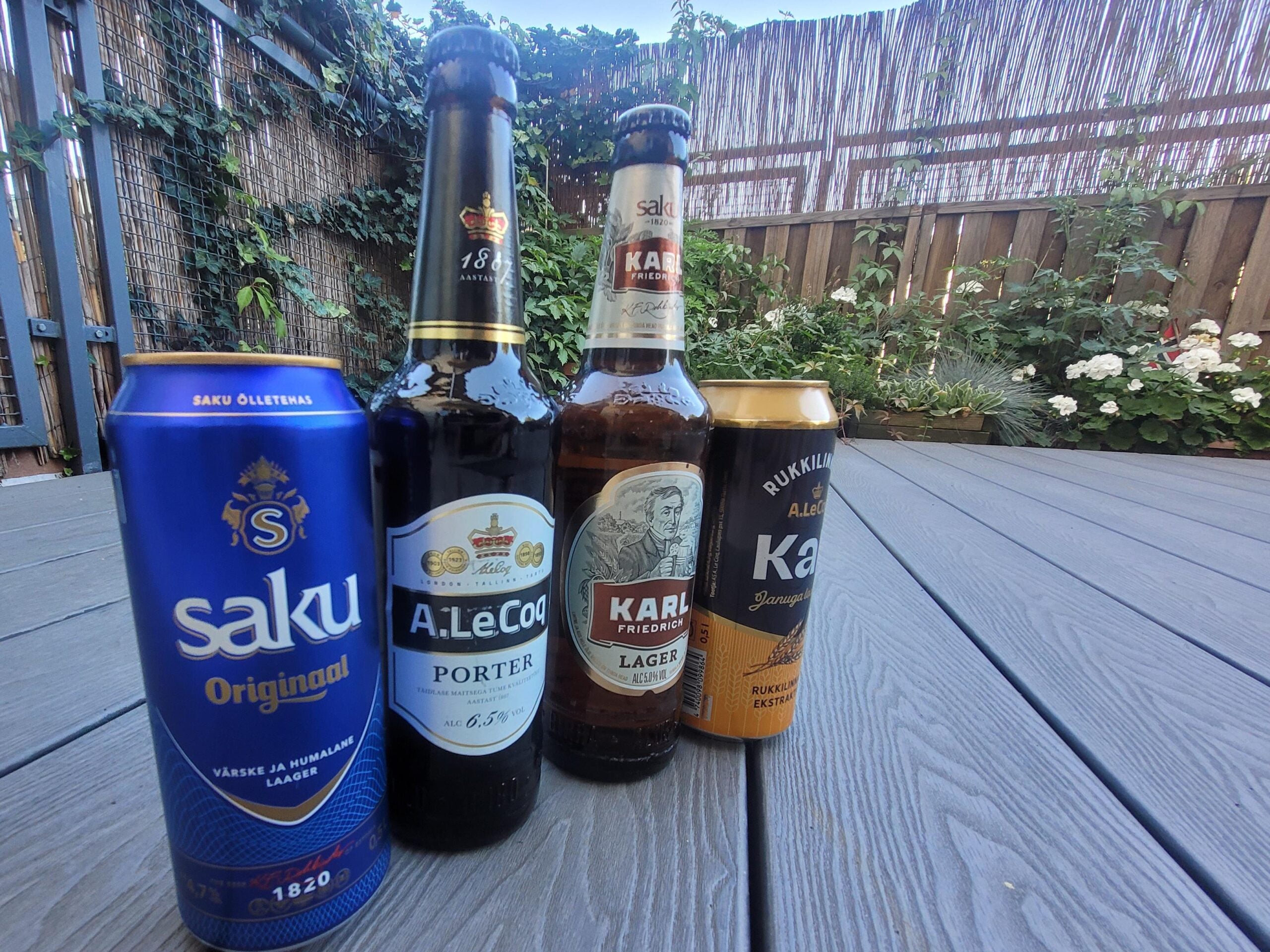 Four different Estonian beer bottles and cans are lined up on a wooden deck outside, with plants and a wooden fence in the background. The labels include Saku Original, A. Le Coq Porter, and two Karl Friedrich beers.