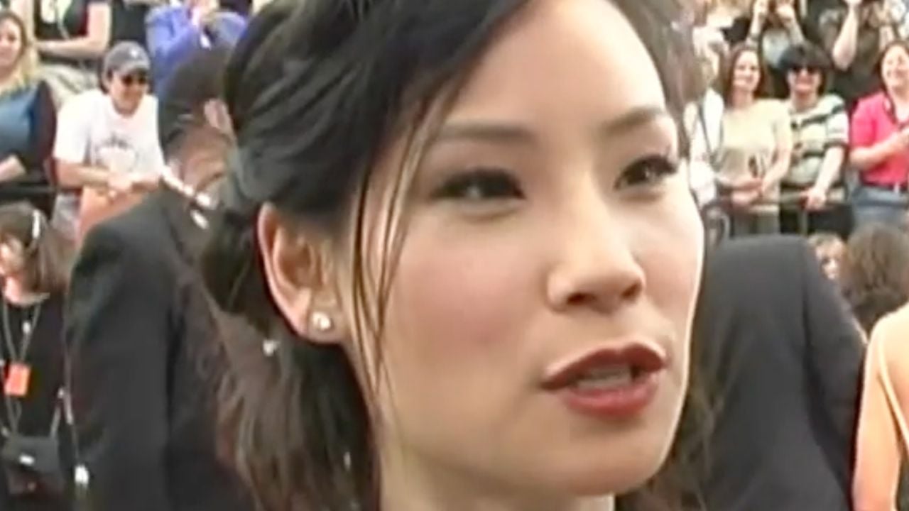 A woman with dark hair and earrings is speaking outdoors, surrounded by a crowd of people. The background is blurred, and she appears to be at an event or gathering.
