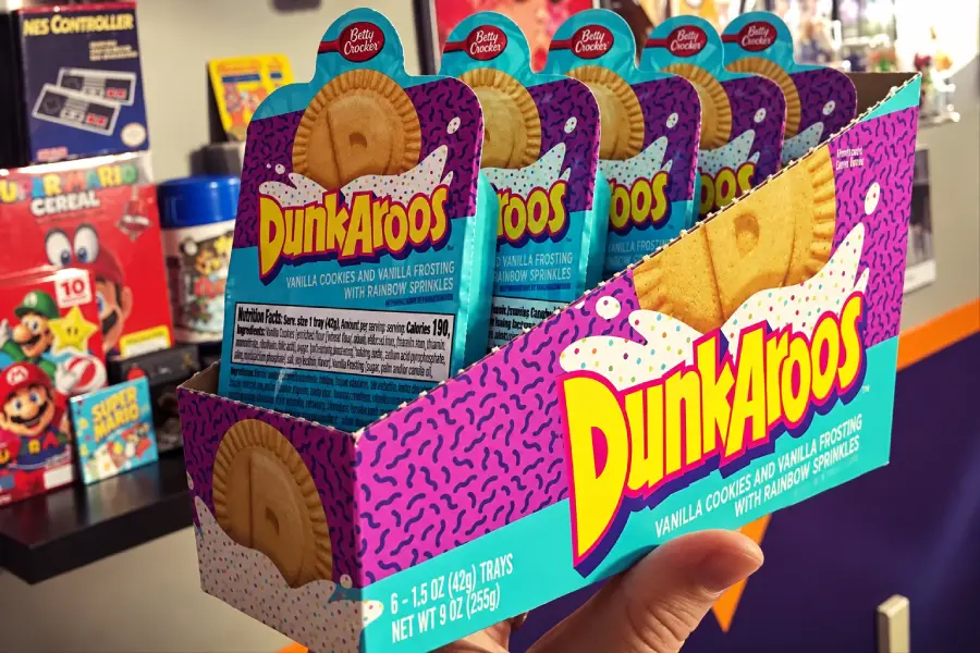 A hand holds a box of Dunkaroos vanilla cookies with rainbow sprinkle frosting. The box is colorful with bold yellow text and shows individual snack trays inside. Retro gaming items are blurred in the background.
