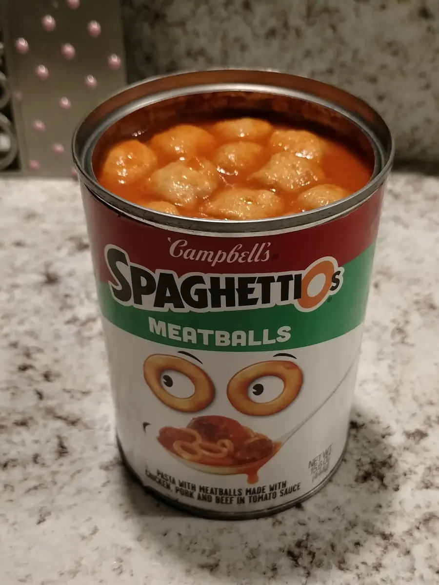 An open can of Campbell’s SpaghettiOs with Meatballs sits on a marble counter, showing meatballs and pasta in tomato sauce inside the can. The label features cartoon spaghetti rings with eyes.