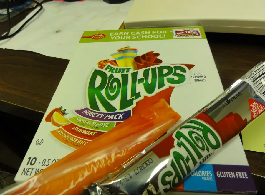 A box of Fruit Roll-Ups Variety Pack is on a table, with an opened orange Fruit Roll-Up in front. The box highlights flavors and a promotion for schools.