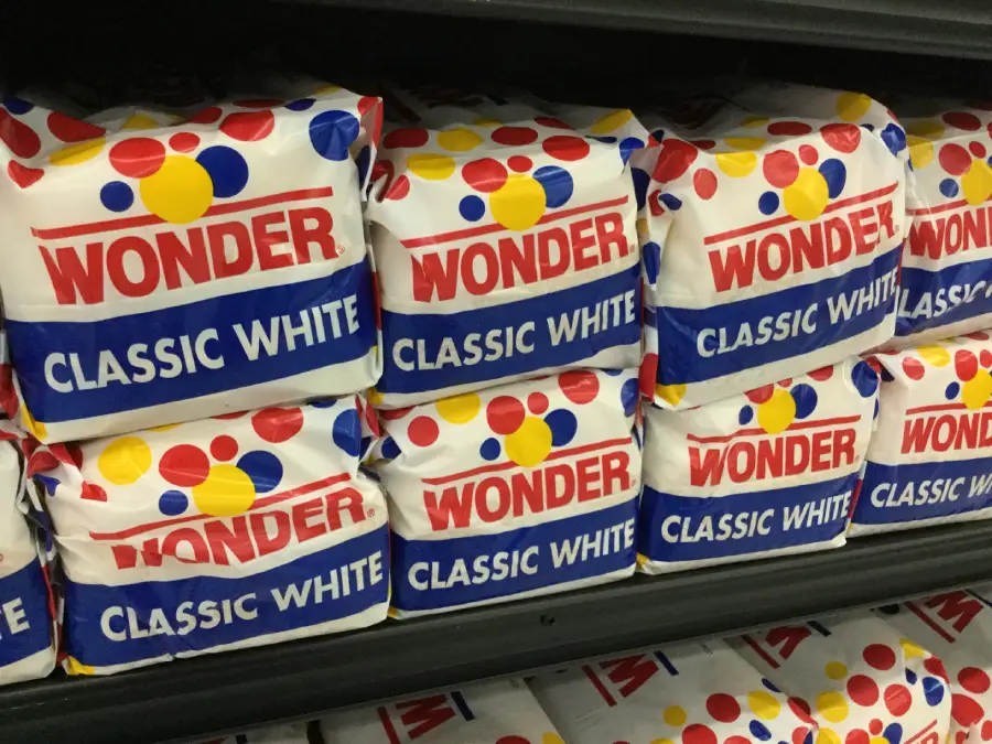 Loaves of Wonder Classic White bread in colorful packaging with red, blue, and yellow dots, stacked on shelves in a grocery store.