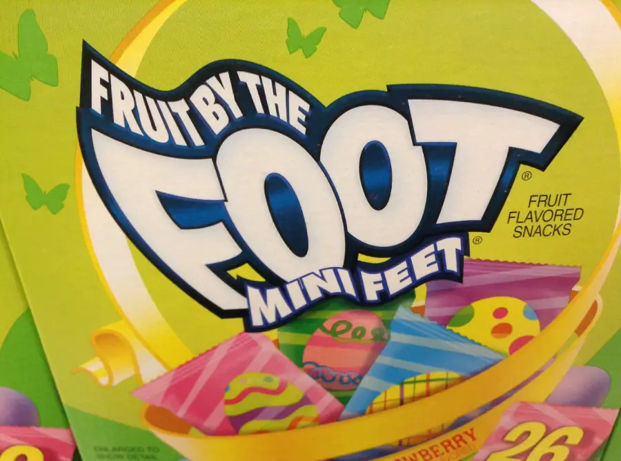 Close-up of a Fruit by the Foot Mini Feet box, showing colorful, patterned snack wrappers and bold white and blue lettering on a green and yellow background with butterfly designs.