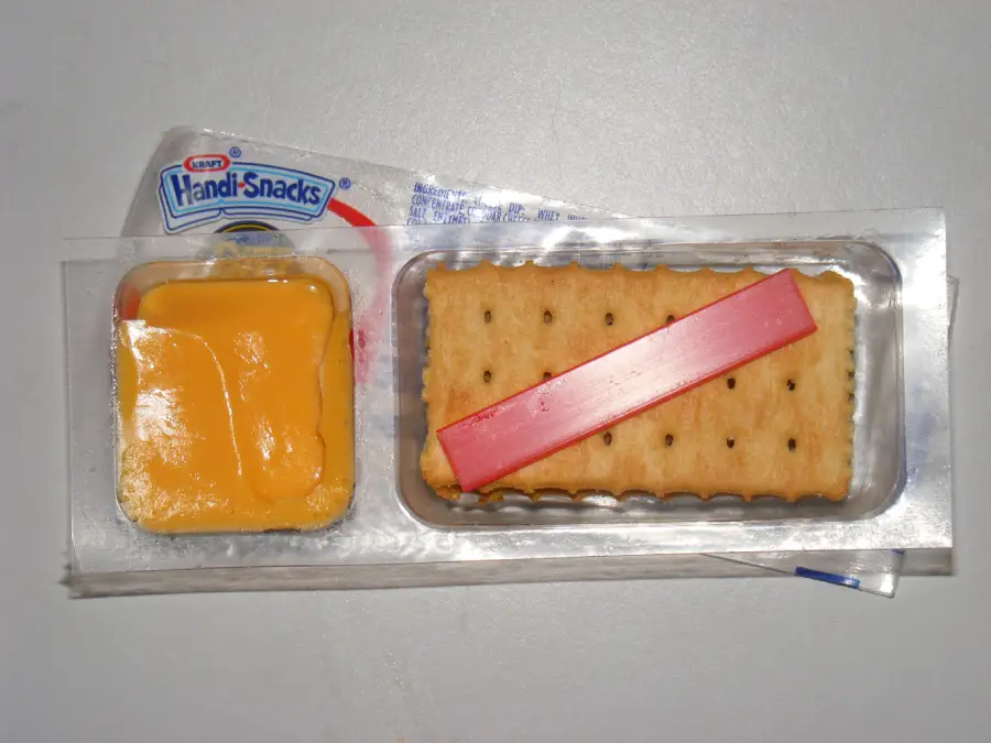 Handi Snacks package with processed cheese on the left, a red cheese spreader on top of stacked crackers on the right, all on a white surface.