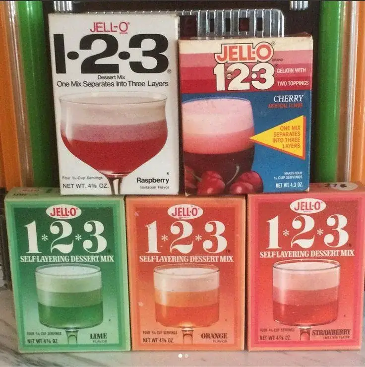 Five vintage Jell-O 1-2-3 dessert mix boxes in raspberry, cherry, lime, orange, and strawberry flavors, displayed upright in two rows on a striped background. Each box shows dessert images and branding.