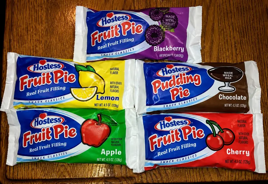 Five Hostess pies on a wooden surface: Blackberry, Lemon, Chocolate Pudding Pie, Apple, and Cherry, each in colorful packaging labeled with fruit images and “Real Fruit Filling” or “Made with Real Cocoa.”