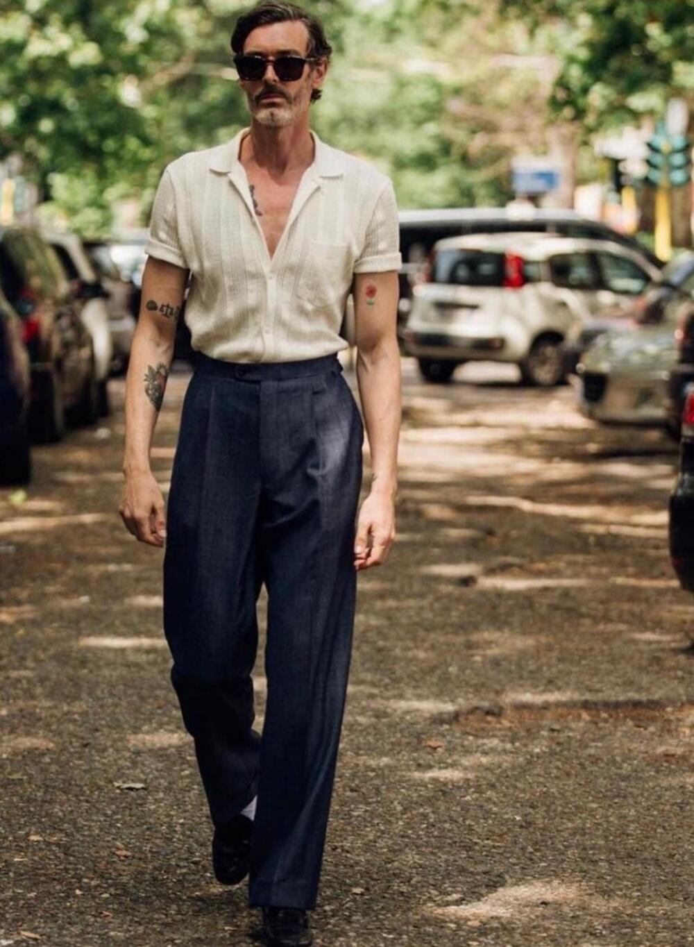 A man with sunglasses and tattoos walks on a tree-lined street wearing a white short-sleeve shirt tucked into high-waisted dark trousers. Cars are parked along both sides of the street in the background.