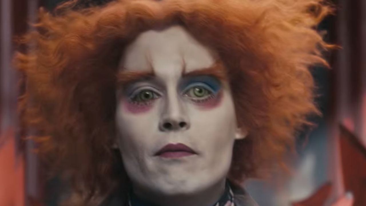 Person with wild orange hair, white face makeup, exaggerated eyebrows, blue and red eyeshadow, and dark lips, wearing a colorful costume, staring directly ahead.