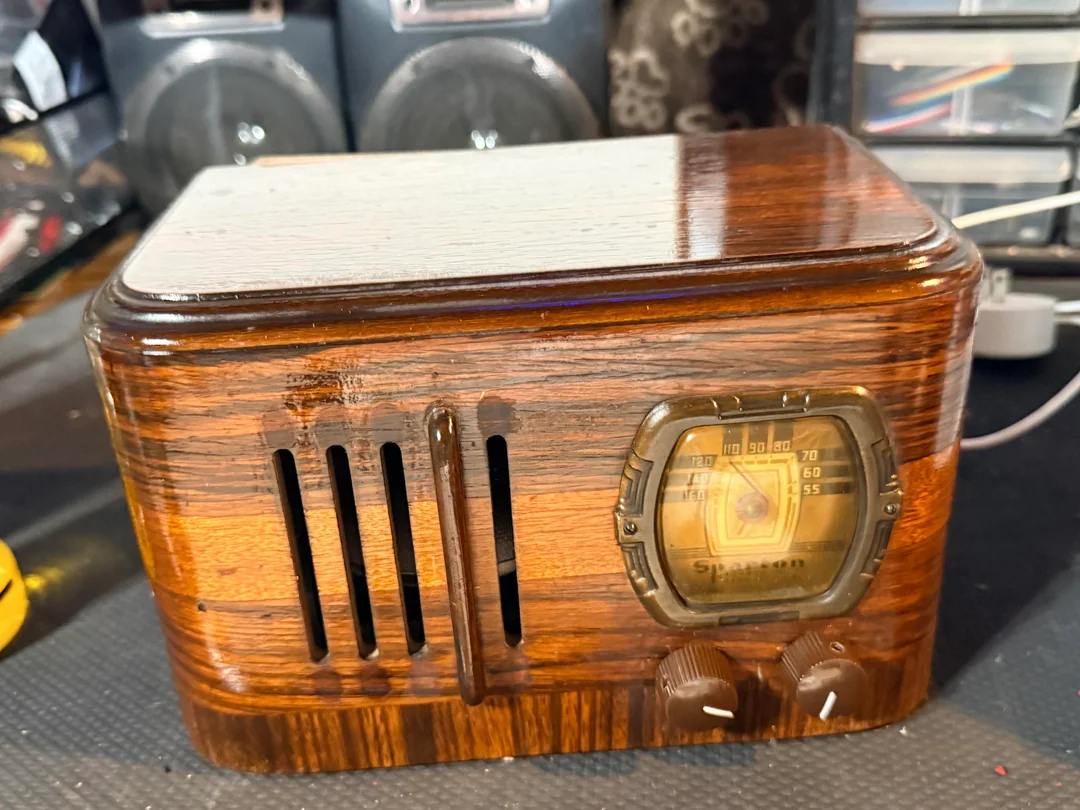 A vintage wooden radio with a rectangular shape, two dials, a small speaker grille, and a glass-covered tuning display sits on a cluttered surface.