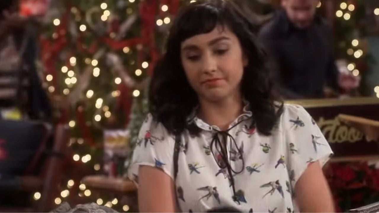 A woman with dark, wavy hair wearing a white blouse with colorful birds stands indoors. In the background, there are Christmas lights and a decorated tree.