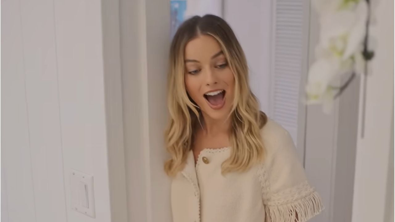 A woman with long blonde hair and a cream-colored top stands indoors, smiling with her mouth open in surprise or excitement.