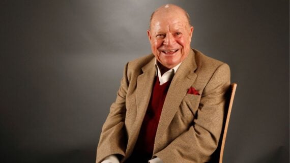 An older man with a bald head, wearing a tan blazer, red sweater, and white shirt, sits smiling on a wooden chair against a dark gray background.