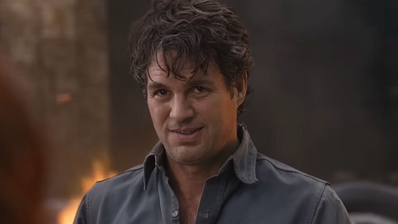 A man with tousled dark hair and a slight smile wears a gray button-up shirt, standing in front of a blurred background with hints of fire and debris.