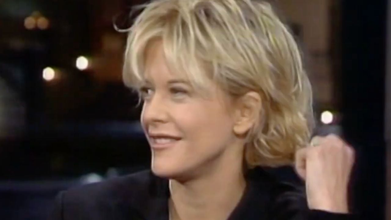 A person with short, blonde hair smiles and looks to the side, wearing a dark jacket. The background is softly blurred with some lights visible.